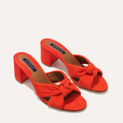 The Carmine Mule - Chili Suede by margaux-development features bright orange open-toe block heels with plush knot details on the front straps and a navy blue Margaux label on the insole, blending comfort and style.