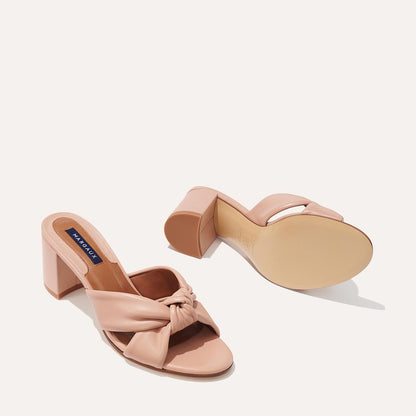 The Carmine Mule - Rose Nappa by margaux-development features blush pink leather, open toes, plush knotted straps, and a block heel. Displayed with one shoe upright and the other showing the sole to highlight comfort and style.