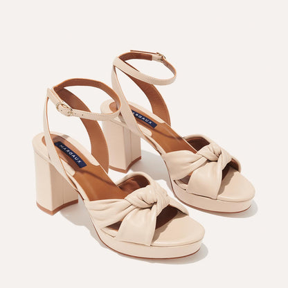 The Carmine Platform - Bone Nappa by margaux-development features beige high-heeled sandals with thick platform block heels, knotted toe straps, and gold-buckle ankle straps for stylish comfort on a light background.
