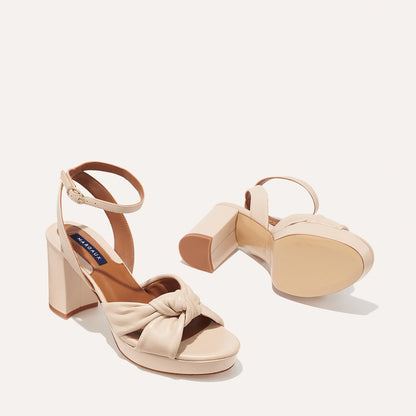 The Carmine Platform - Bone Nappa by margaux-development features beige block heels, ankle straps, and front knotted detailing for style and comfort. One shoe stands upright while the other lays on its side to display the sole.