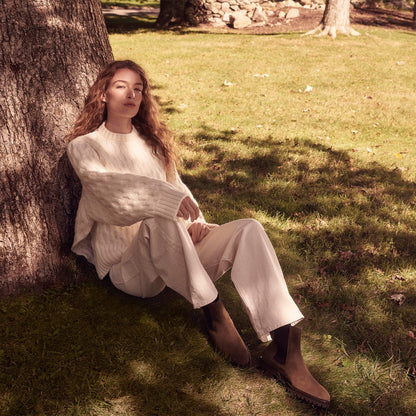 A person with long wavy hair, dressed in a cream sweater and white pants, sits on grass leaning against a tree under the sun, wearing Margaux's The Chelsea Boot in Moss Suede.