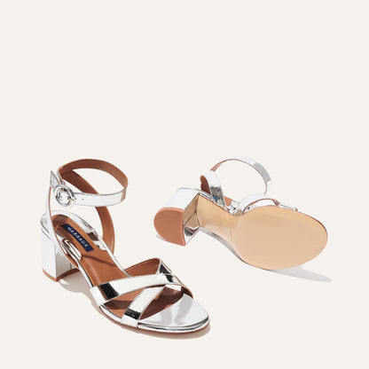 The City Sandal - Silver Mirror by margaux-development features shiny silver open-toe heels with crisscross straps, ankle buckles, and a cushioned insole for comfort. One sandal stands upright; the other is shown sole-up.