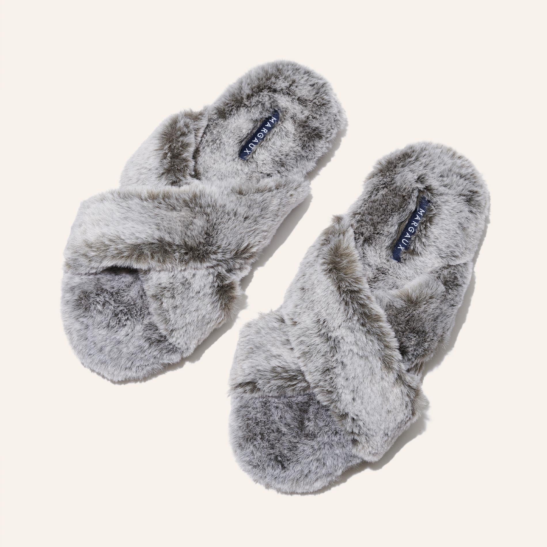 The Cozy Slipper - Grey by Margaux features a soft, fluffy texture and cozy x-strap design. This stylish faux fur slipper is displayed on a plain light background.