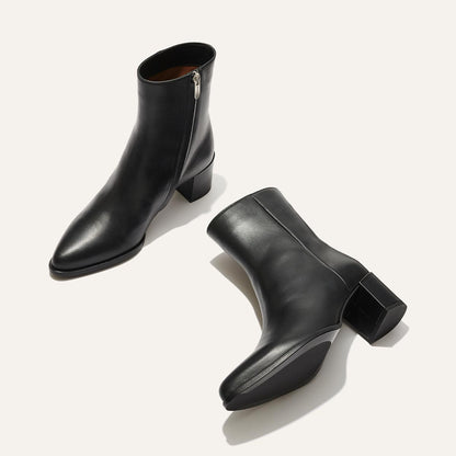 A pair of black leather ankle boots with side zippers and block heels displayed on a light background, one boot upright and the other lying on its side.