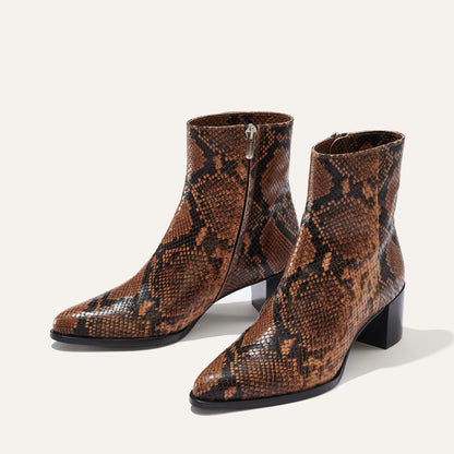 The Downtown Boot First Edition by Margaux features a brown and black python-embossed pattern, mid heels, pointed toes, and side zippers—western-inspired ankle boots shown on a plain light background.