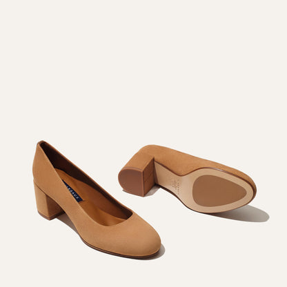 The Heel - Cacao Suede by Margaux: One elegant tan suede block heel pump stands upright, highlighting the rounded toe and side, while the other rests on its side to display the sole and stylish block heel against a plain light background.