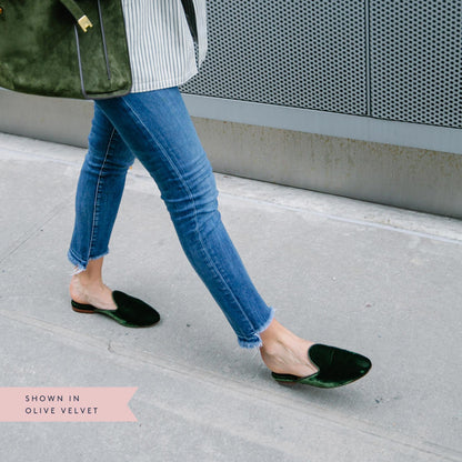 A person walks on a city sidewalk wearing blue jeans, an olive green bag, and The Loafer Mule - Olive Velvet by Margaux. Text on the image reads, Shown in olive velvet.