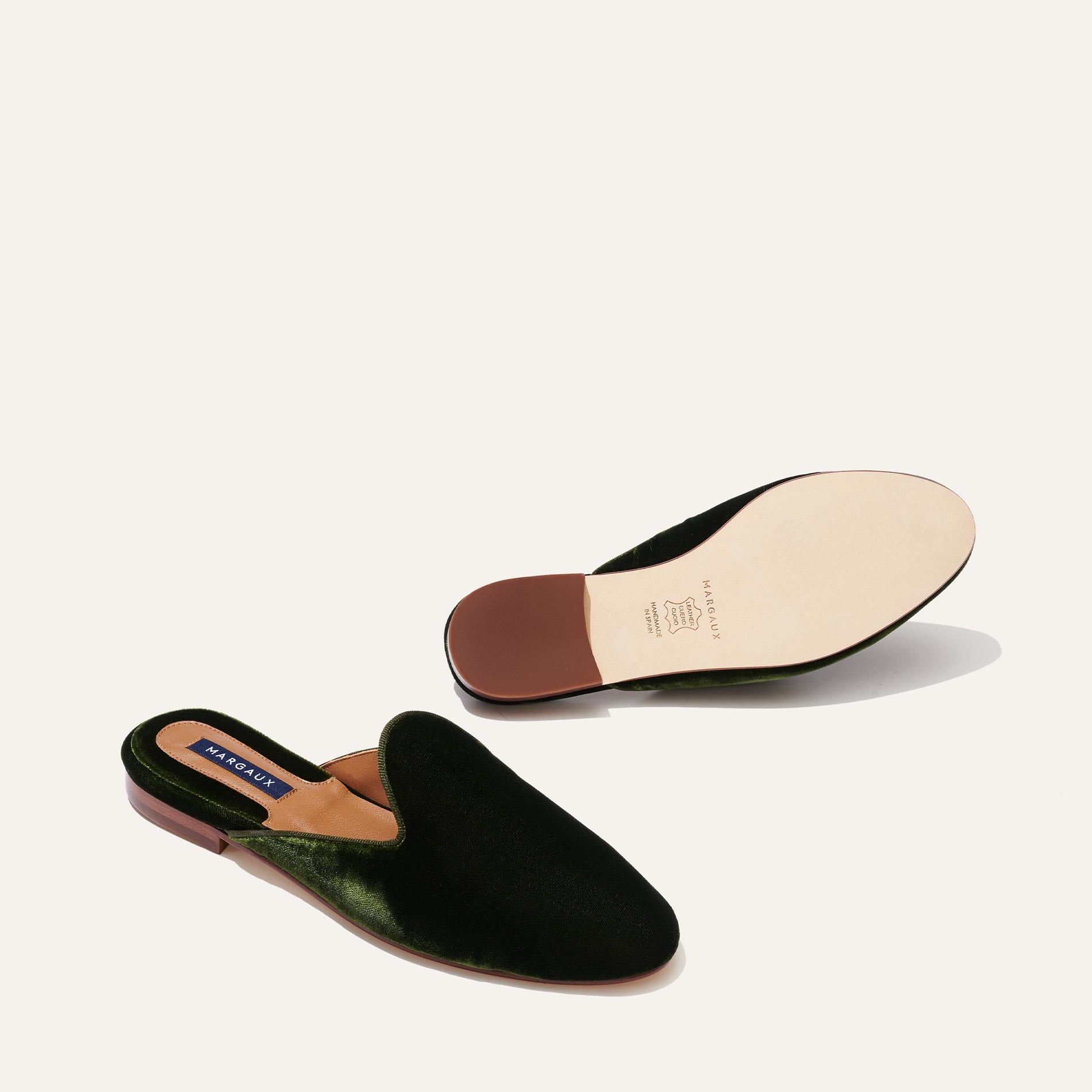 The Loafer Mule - Olive Velvet by Margaux, featuring dark green velvet, almond toes, and tan leather soles; one shoe is shown from above while the other displays the smooth sole and brown heel.