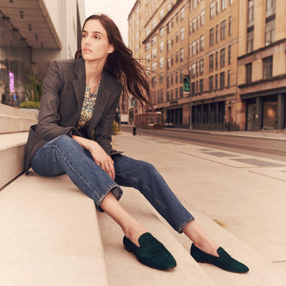 A woman with long brown hair, dressed in a gray blazer, floral top, blue jeans, and Margaux's The Penny - Emerald Suede loafers, sits on city steps among tall buildings, gazing thoughtfully into the distance.