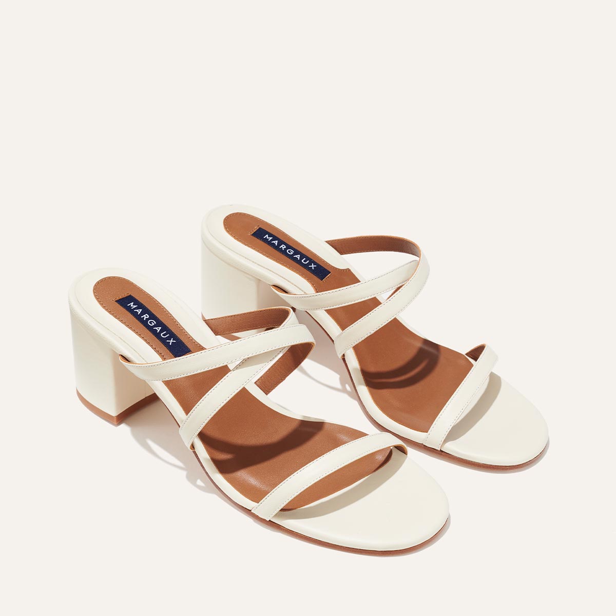 The Perry Sandal - Ivory Nappa by margaux-development showcases stylish ivory Nappa leather block heels with two crisscross straps and brown insoles, set against a plain light background.
