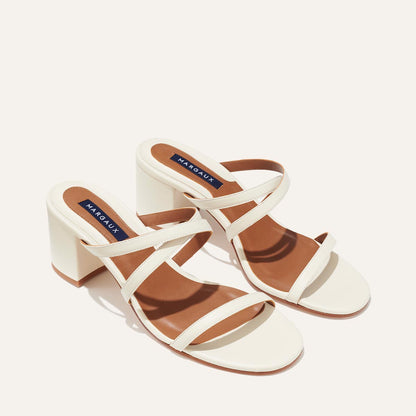 The Perry Sandal - Ivory Nappa by margaux-development showcases stylish ivory Nappa leather block heels with two crisscross straps and brown insoles, set against a plain light background.