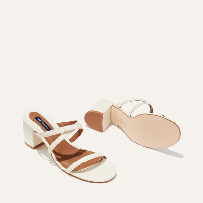 The Perry Sandal - Ivory Nappa by margaux-development features soft Nappa leather, two thin straps, open toes, and a chic block heel. One sandal stands upright, the other lies on its side displaying the sole and heel.