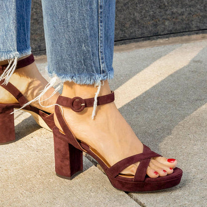 A person wearing frayed-hem blue jeans and margaux-development’s The Platform Sandal - Bordeaux Suede stands on a concrete sidewalk. Their red-painted toenails complement the chic ankle-strap sandals for an elevated look.