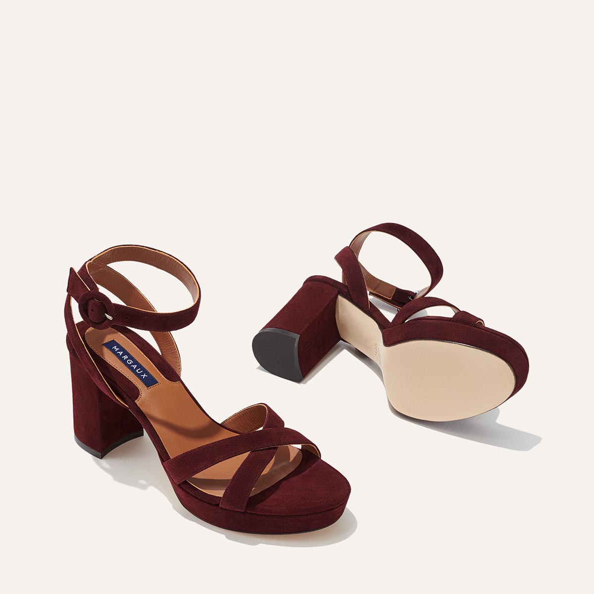 The Platform Sandal - Bordeaux Suede by margaux-development features thick heels, ankle straps, and crisscross toe straps for a comfortable, elevated look—one shoe upright and one on its side against a light background.