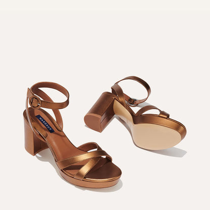 The Platform Sandal - Bronze Nappa by margaux-development features metallic bronze, platform soles, ankle straps, and block heels. One shoe stands upright while the other lies on its side to show the sole.