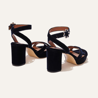 The Platform Sandal by margaux-development in midnight blue velvet features thick block heels, crisscross and ankle straps with floral buckles, showcased on a light background.