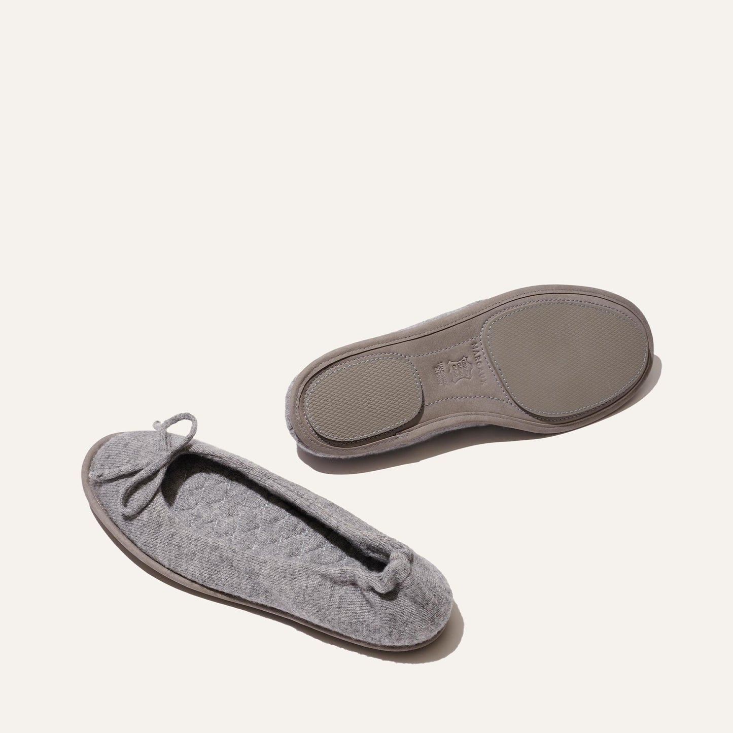 A pair of Margaux's The Slipper - Heather, gray quilted ballet flat luxury slippers with plush innersoles—one displays a small bow, the other the textured rubber sole—set on a plain off-white background.
