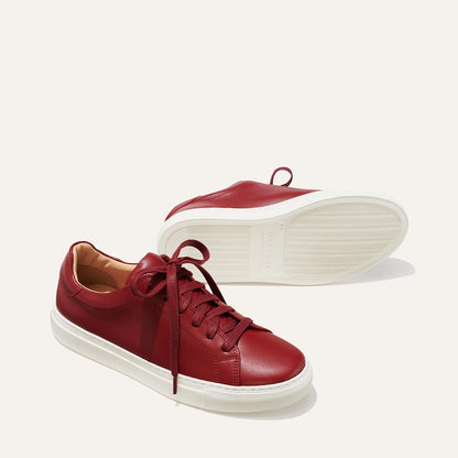 The Sneaker - Burgundy Calf by margaux-development: a pair of burgundy leather sneakers with white rubber soles. One stands upright with laces tied; the other lies on its side, displaying the textured sole against a plain, light background.