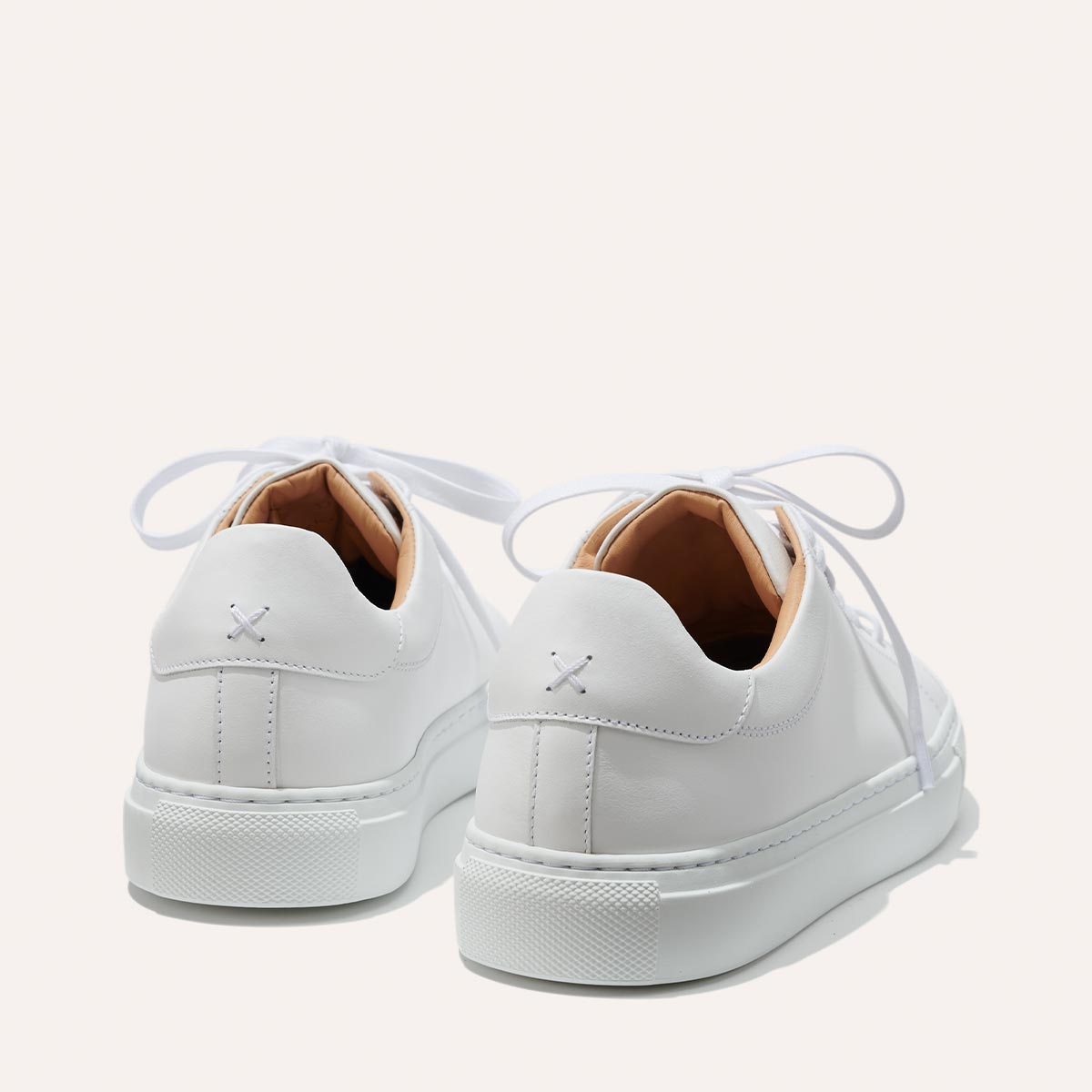 The Sneaker - White Calf by margaux-development is a white leather sneaker with white laces, tan inner lining, and an X-shaped stitch on the heel. Its minimalist design is shown from the back and slightly to the side.