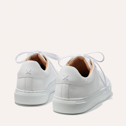 The Sneaker - White Calf by margaux-development is a white leather sneaker with white laces, tan inner lining, and an X-shaped stitch on the heel. Its minimalist design is shown from the back and slightly to the side.