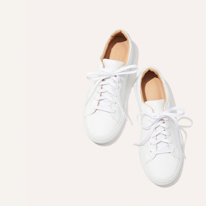 A pair of margaux-development's The Sneaker - White Calf, featuring white leather and a light tan inner lining, is shown side by side with loosely tied laces on a neutral background.