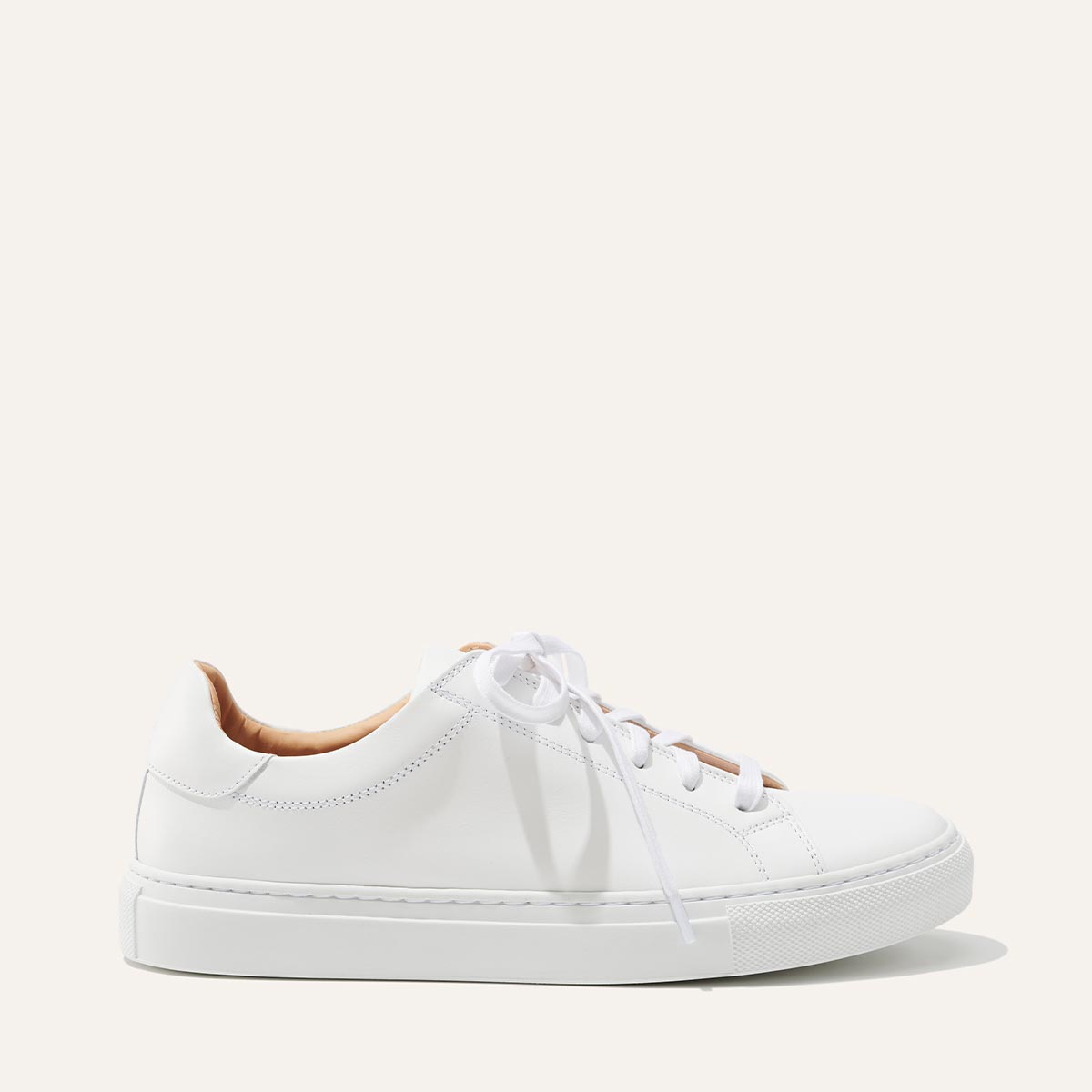 The Sneaker - White Calf by margaux-development is a white low-top sneaker with white laces and a thick rubber sole, shown in profile against a plain, light background.