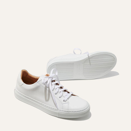 The Sneaker - White Calf by margaux-development features a white low-top design, white laces, tan inner lining, and minimalist style. One sneaker stands upright while the other is laid on its side to display the sole.