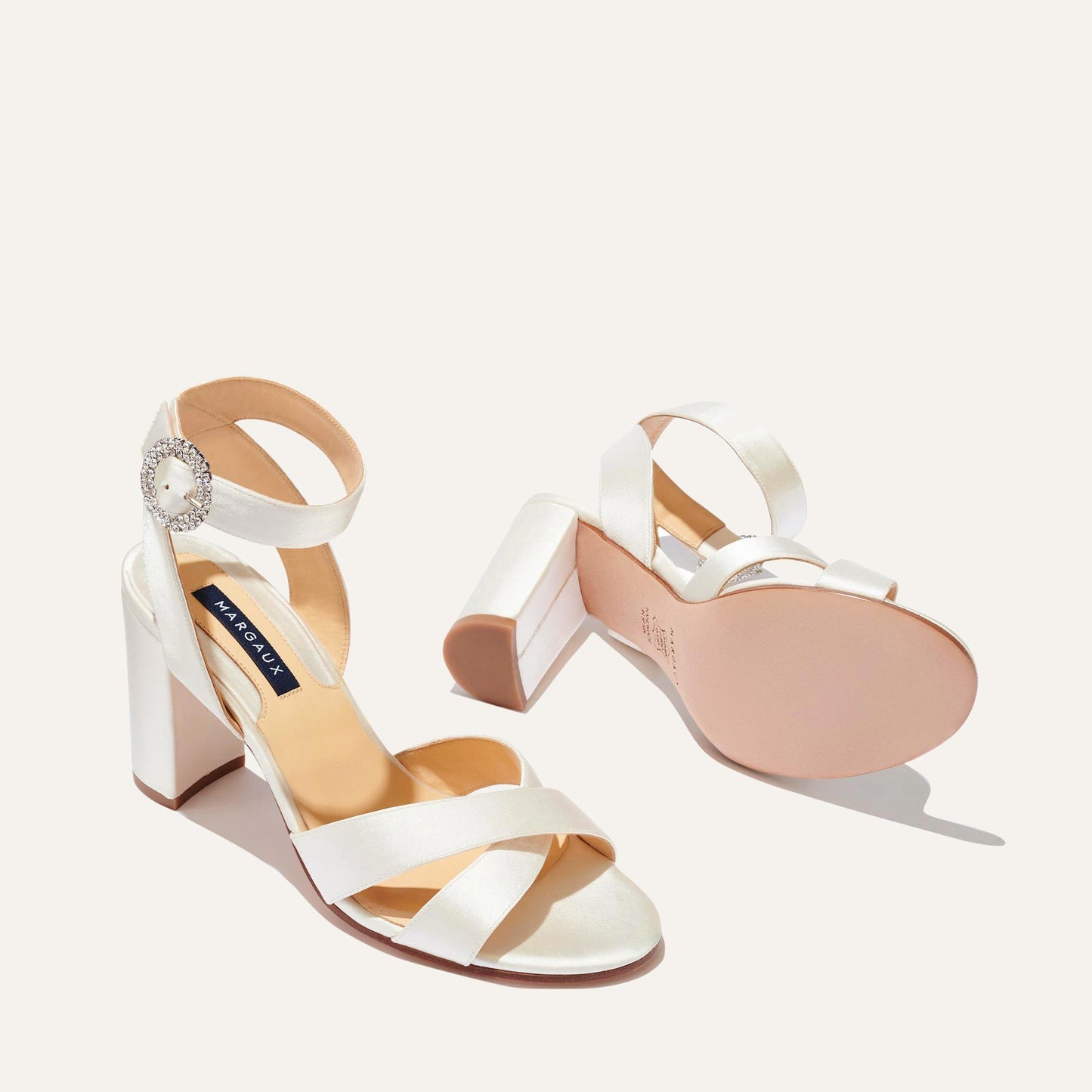The Uptown Sandal - Ivory Satin with Crystal Buckle by margaux-development features elegant crisscross and ankle straps, a sparkling rhinestone buckle, beige sole, and block heel, shown on a light background.