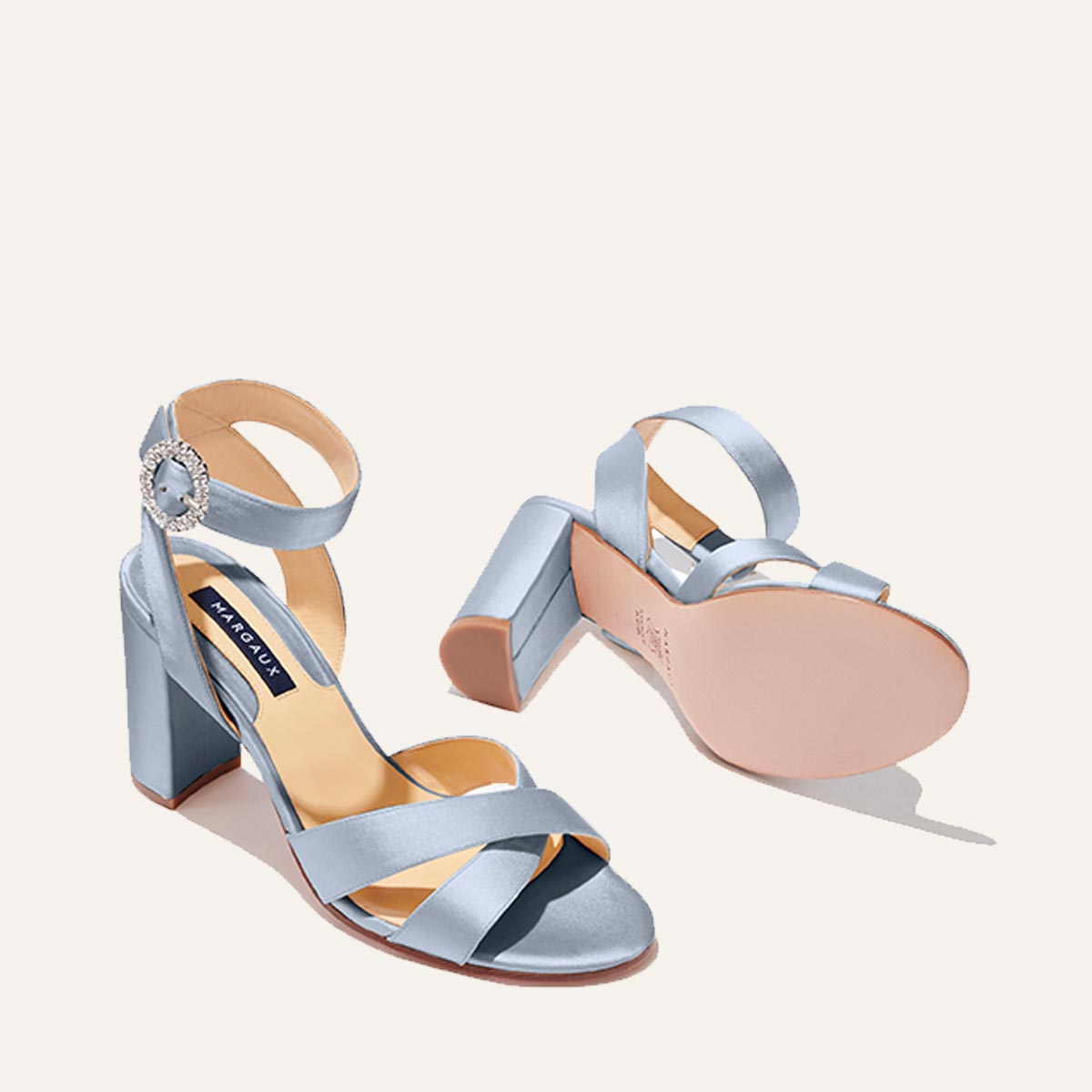 The Uptown Sandal - Powder Satin with Crystal Buckle by margaux-development features light blue satin block heels, ankle straps, and rhinestone buckles—one stands upright, the other lies on its side to reveal the sole. Designed for all-day comfort.
